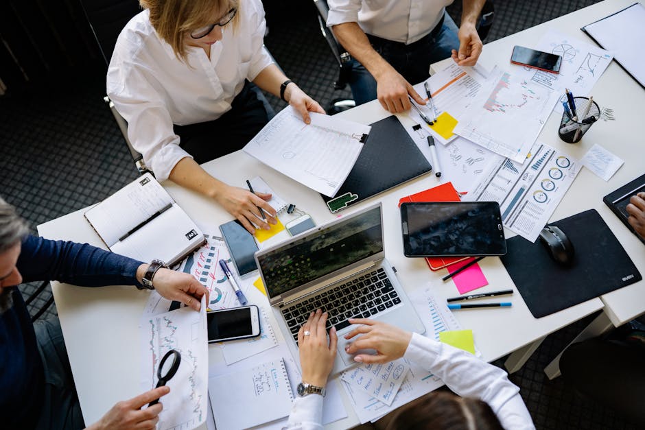 A group of colleagues working around a table, reviewing documents and using laptops and tablets.